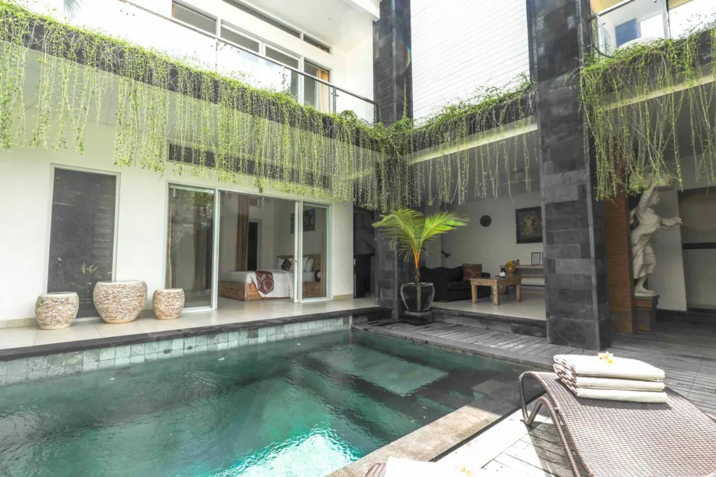 Serene private infinity pool in a modern Balinese villa, with lush hanging vines, black stone pillars, lounge chairs, and tropical plants. - Bali Villas