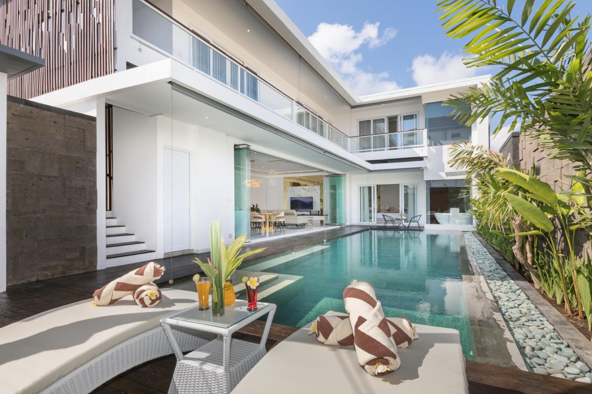 Modern luxury Bali villa with infinity pool, outdoor loungers, tropical plants, and glass-walled living area. - Bali Villas