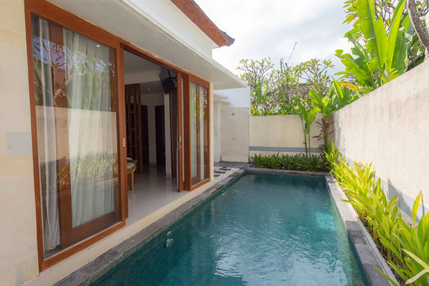 Serene private infinity pool in a Balinese villa, with open wooden sliding doors to a modern interior and lush tropical plants. - Bali Villas
