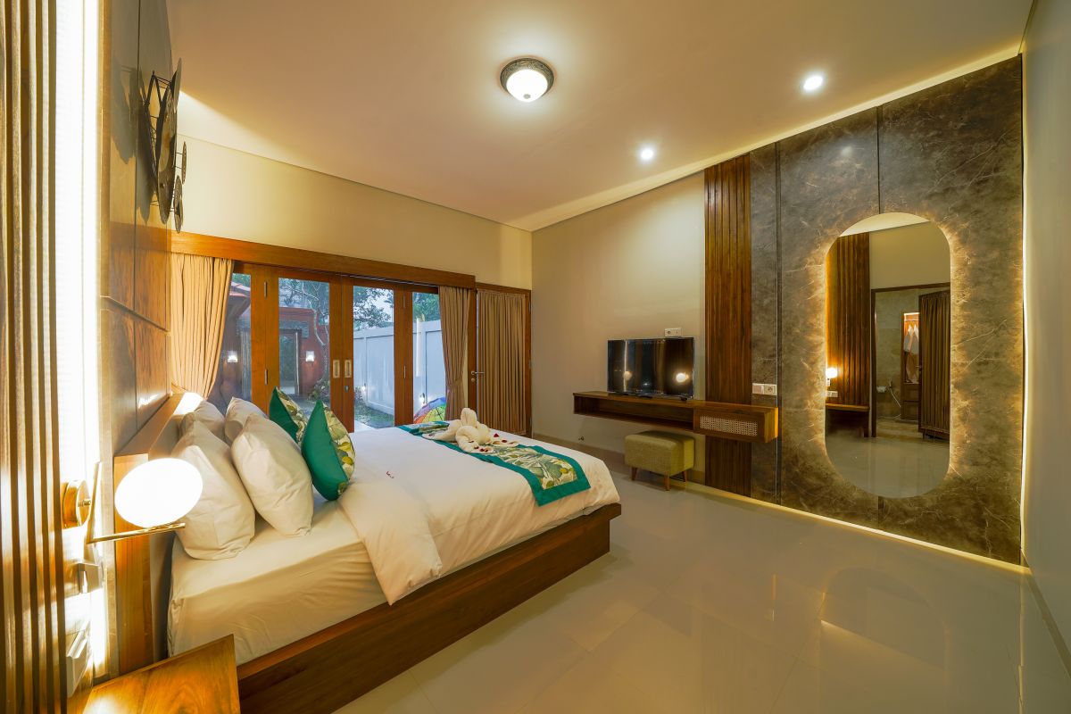 Luxurious Balinese bedroom with wooden king bed, green accents, TV, and sliding doors to garden view. - Bali Villas