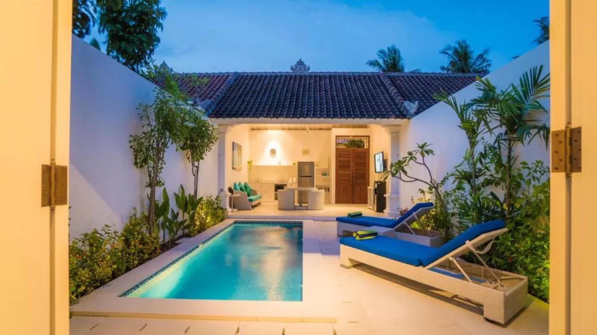 Modern tropical villa interior with blue sofa, open kitchen, wall-mounted TV, and wooden doors to outdoor patio. - Bali Villas