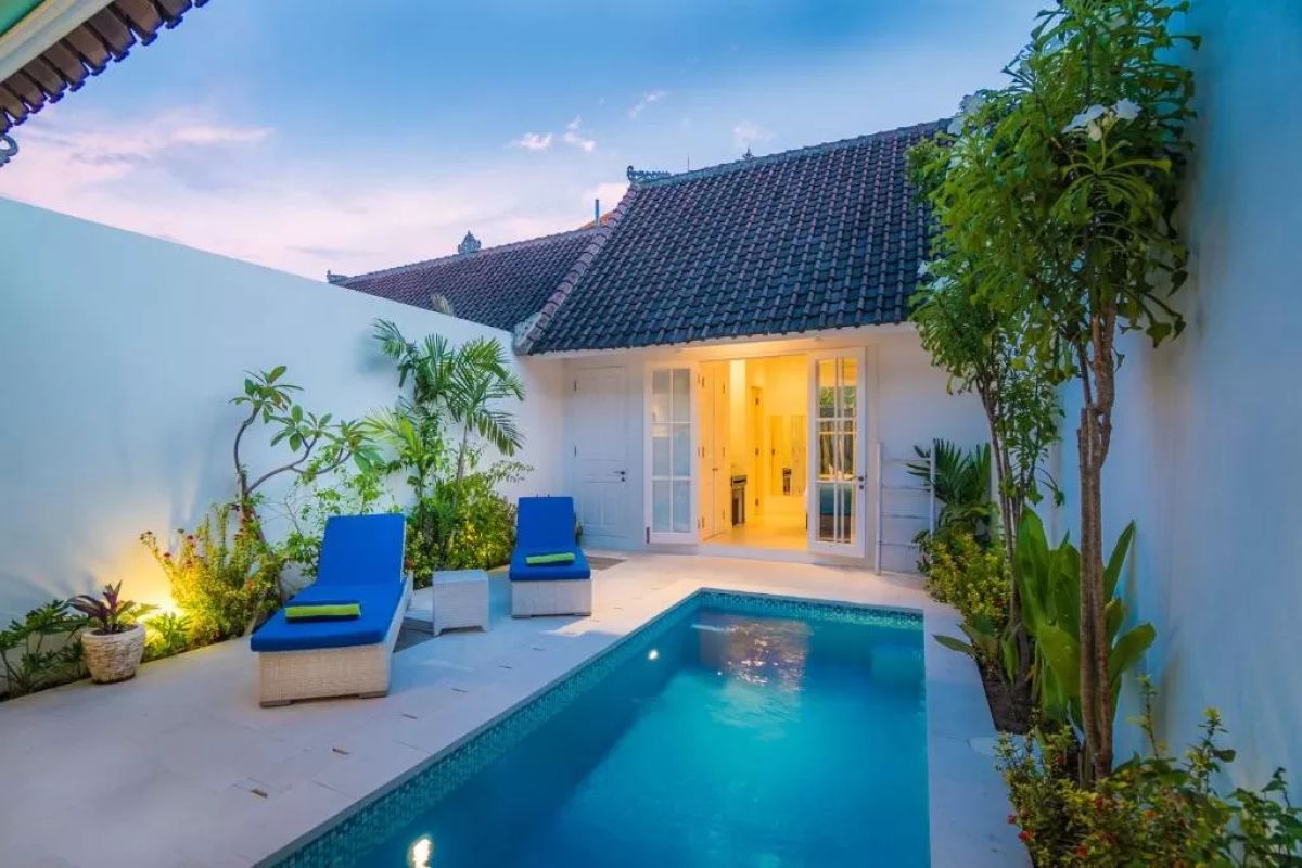 Modern tropical villa interior with blue sofa, open kitchen, wall-mounted TV, and wooden doors to outdoor patio. - Bali Villas