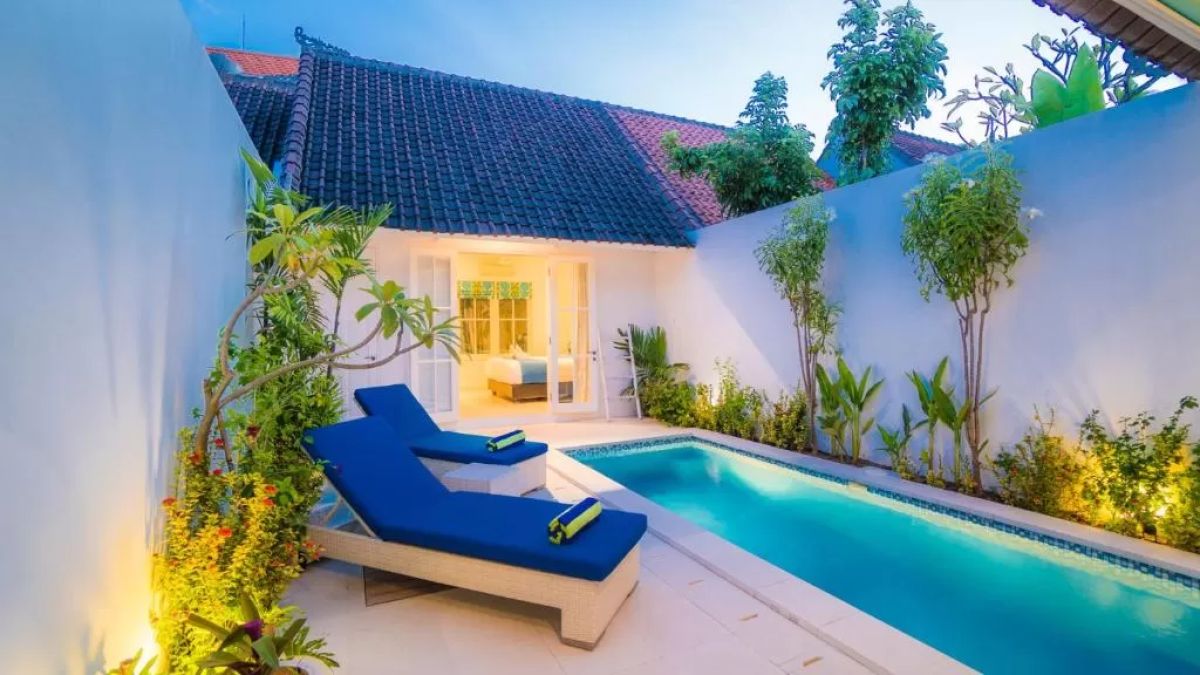 Modern tropical villa interior with blue sofa, open kitchen, wall-mounted TV, and wooden doors to outdoor patio. - Bali Villas