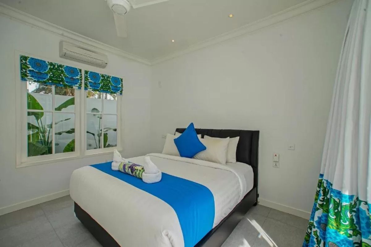Modern tropical villa interior with blue sofa, open kitchen, wall-mounted TV, and wooden doors to outdoor patio. - Bali Villas