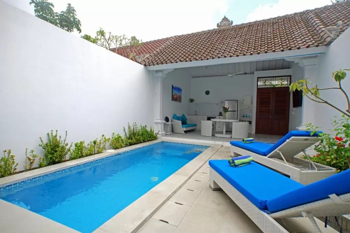 Modern tropical villa interior with blue sofa, open kitchen, wall-mounted TV, and wooden doors to outdoor patio. - Bali Villas