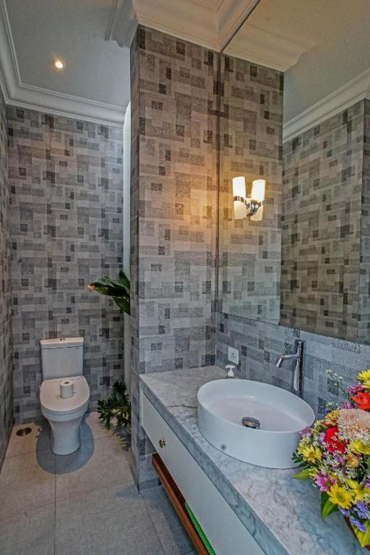 Modern gray-tiled bathroom featuring a white sink vanity, mirror, toilet, potted plants, and decorative window. - Bali Villas