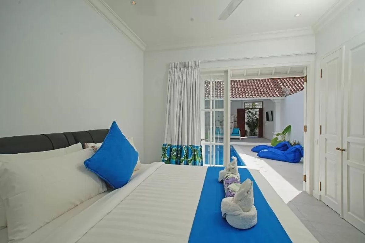 Modern tropical villa interior with blue sofa, open kitchen, wall-mounted TV, and wooden doors to outdoor patio. - Bali Villas
