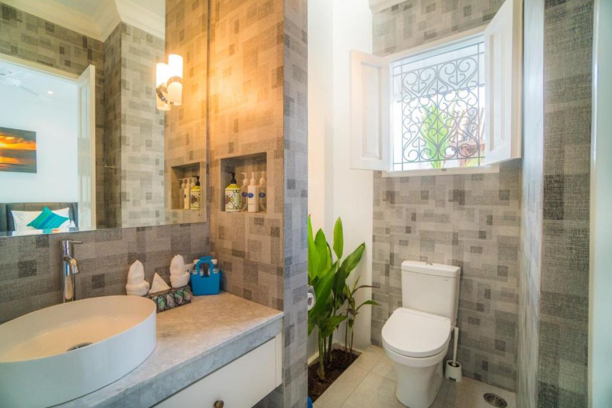 Modern gray-tiled bathroom featuring a white sink vanity, mirror, toilet, potted plants, and decorative window. - Bali Villas