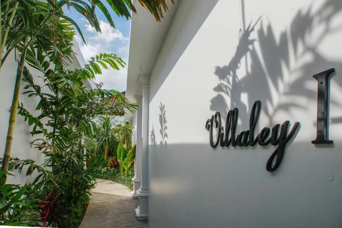 Modern tropical villa interior with blue sofa, open kitchen, wall-mounted TV, and wooden doors to outdoor patio. - Bali Villas