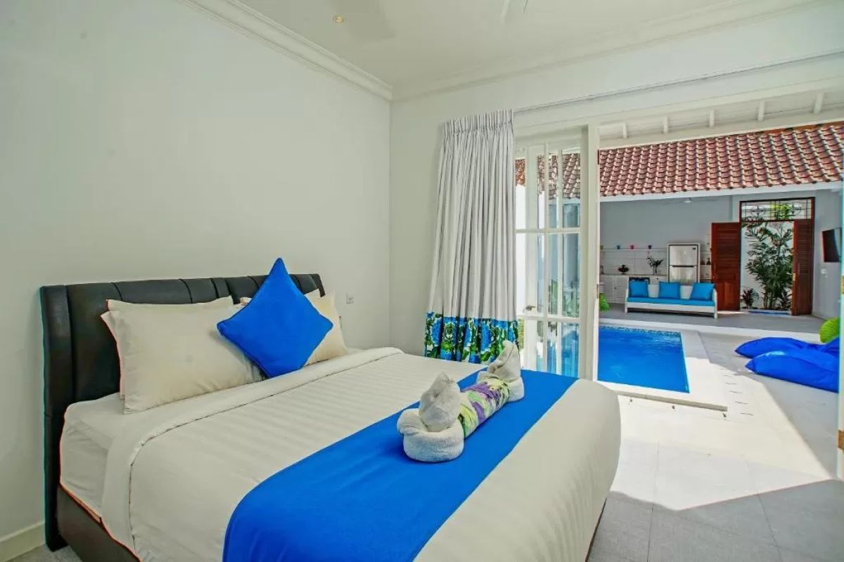 Modern tropical villa interior with blue sofa, open kitchen, wall-mounted TV, and wooden doors to outdoor patio. - Bali Villas