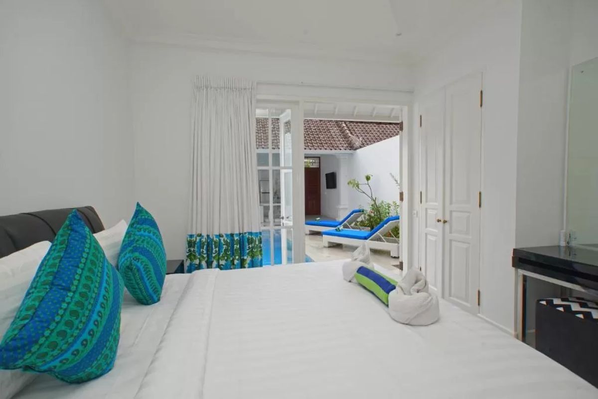 Modern tropical villa interior with blue sofa, open kitchen, wall-mounted TV, and wooden doors to outdoor patio. - Bali Villas