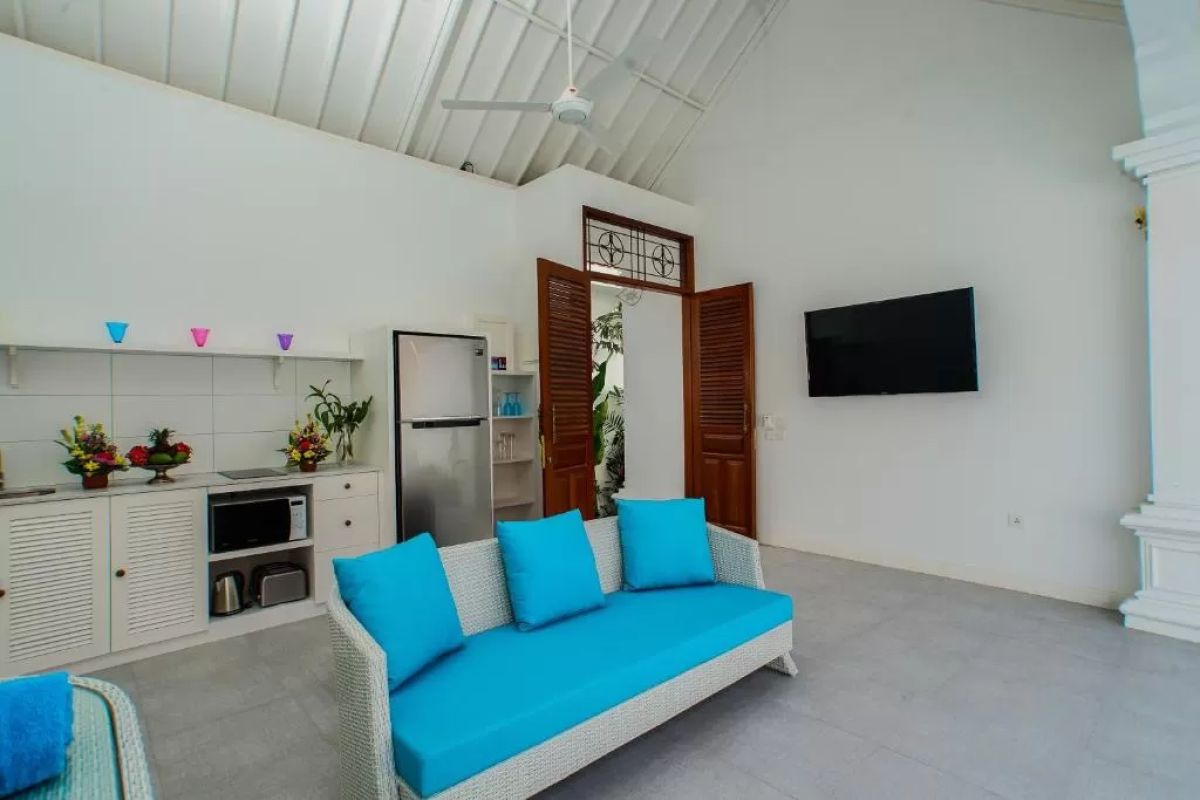 Modern tropical villa interior with blue sofa, open kitchen, wall-mounted TV, and wooden doors to outdoor patio. - Bali Villas