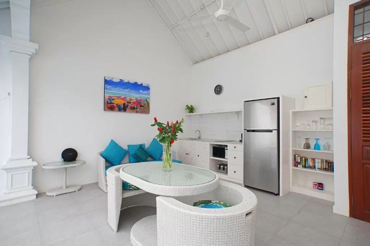 Modern tropical villa interior with blue sofa, open kitchen, wall-mounted TV, and wooden doors to outdoor patio. - Bali Villas