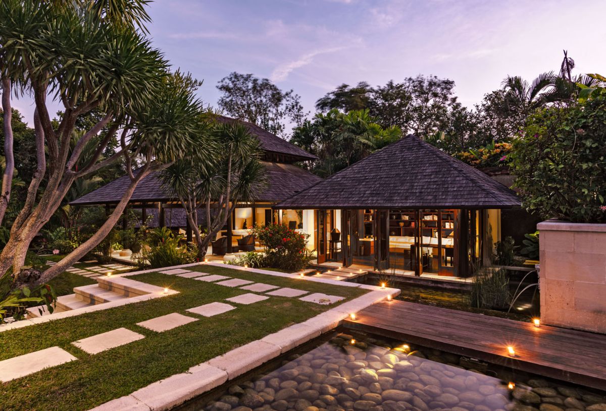 Luxurious Balinese villa at dusk with thatched roofs, tropical palms, stone pathways, and illuminated reflecting pond. - Bali Villas