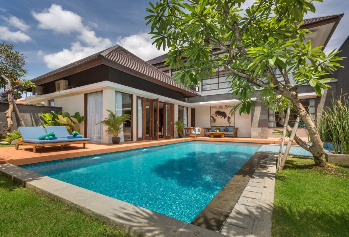 Modern tropical villa with infinity-edge pool, wooden deck lounge, and lush palm trees under a sunny sky. - Bali Villas