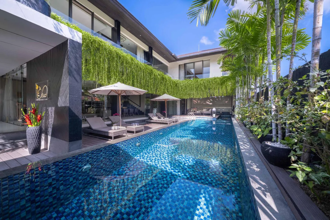 Luxury tropical infinity pool at a modern villa, surrounded by palm trees, sun loungers, and lush vertical gardens under a blue sky. - Bali Villas