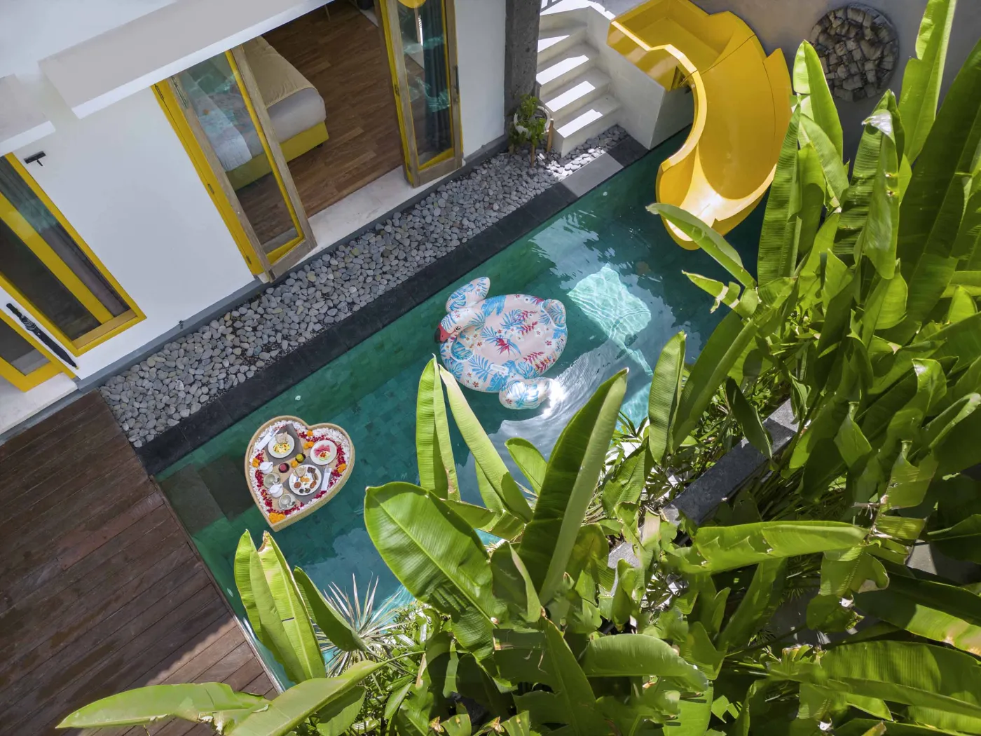 Modern Balinese villa bedroom with yellow accents, open sliding doors to a private turquoise pool, and lush tropical plants on a wooden deck. - Bali Villas