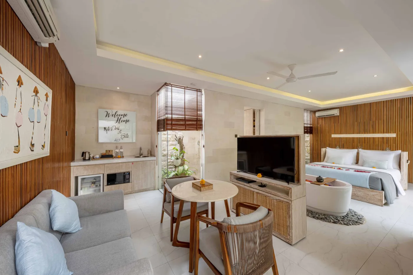 Cozy Balinese villa lounge with wooden paneled walls, gray sofa and chairs around a marble table, abstract wall art, and lush garden window view. - Bali Villas