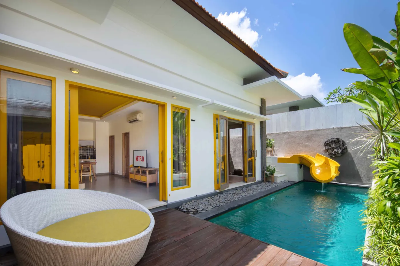 Modern Balinese villa bedroom with yellow accents, open sliding doors to a private turquoise pool, and lush tropical plants on a wooden deck. - Bali Villas