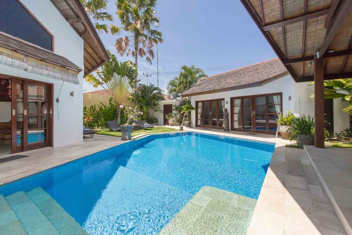 Luxurious Balinese villa pool with clear blue water, surrounded by tropical palms, plants, and shaded verandas. - Bali Villas