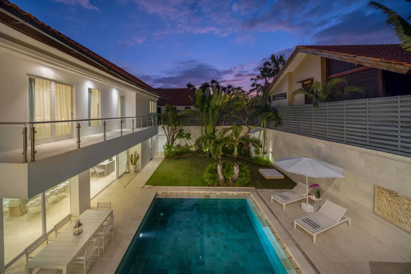 Aerial view of a luxurious modern Bali villa at dusk, featuring an infinity pool, tropical garden, outdoor loungers, and sunset sky. - Bali Villas