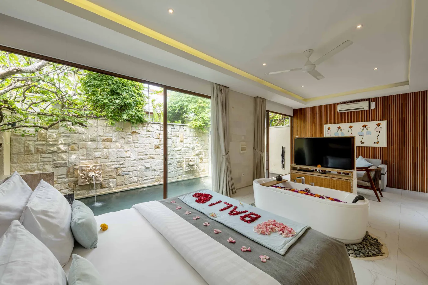 Cozy Balinese villa lounge with wooden paneled walls, gray sofa and chairs around a marble table, abstract wall art, and lush garden window view. - Bali Villas