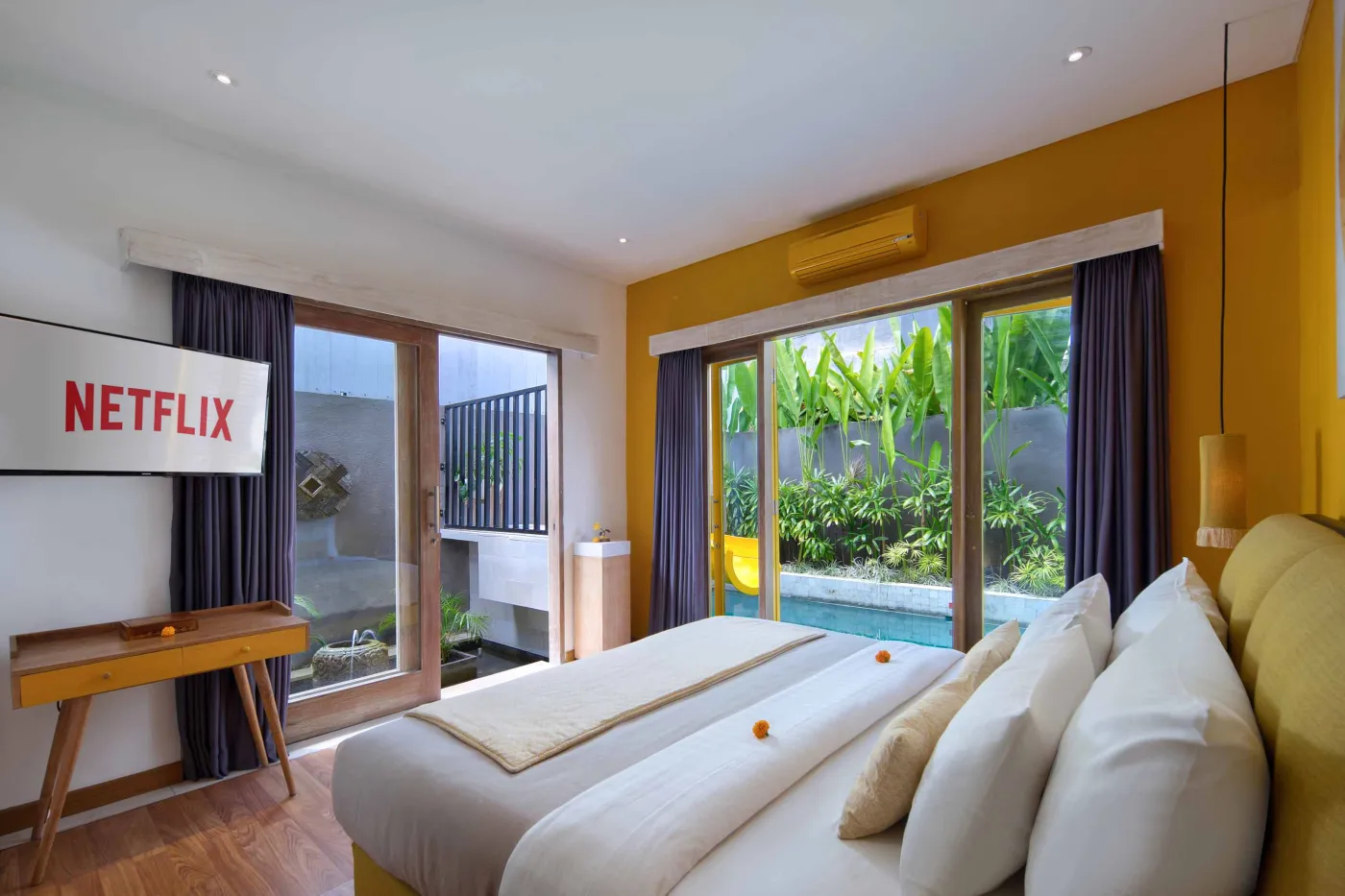 Modern Balinese villa bedroom with yellow accents, open sliding doors to a private turquoise pool, and lush tropical plants on a wooden deck. - Bali Villas