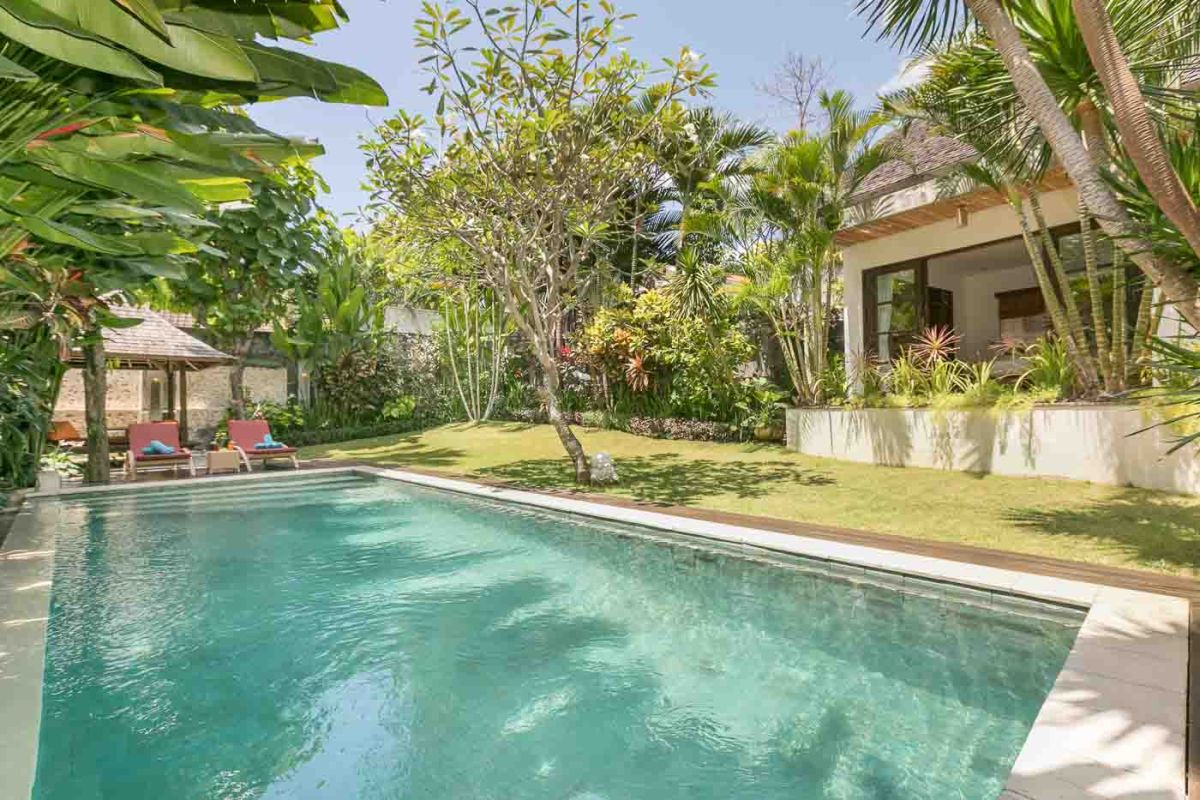 Lush tropical poolside villa in Bali, with turquoise water, palm trees, lounge chairs, and modern thatched architecture. - Bali Villas