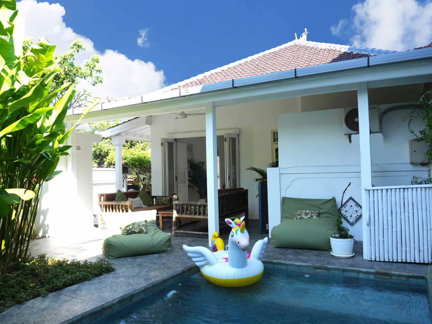Tropical villa pool with yellow unicorn inflatable float, lush green plants, and outdoor lounge seating under blue sky. - Bali Villas