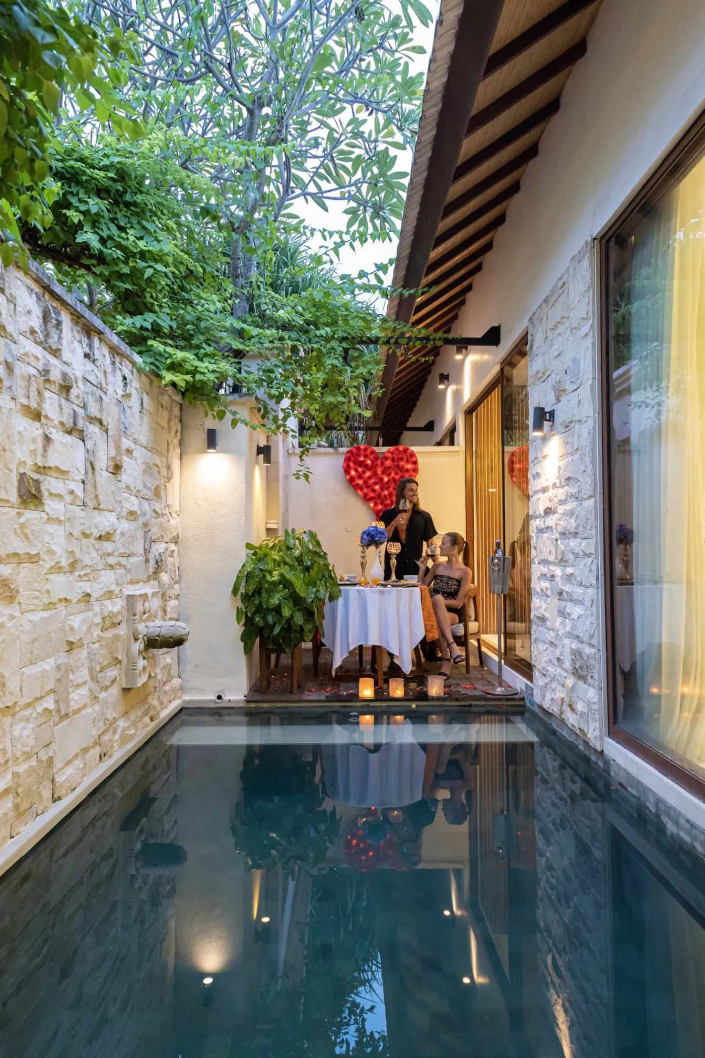 Cozy Balinese villa lounge with wooden paneled walls, gray sofa and chairs around a marble table, abstract wall art, and lush garden window view. - Bali Villas