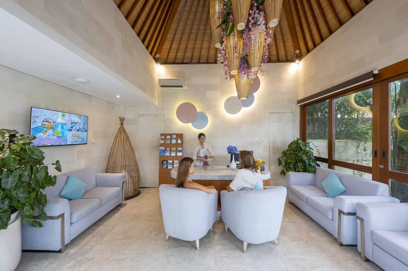 Cozy Balinese villa lounge with wooden paneled walls, gray sofa and chairs around a marble table, abstract wall art, and lush garden window view. - Bali Villas