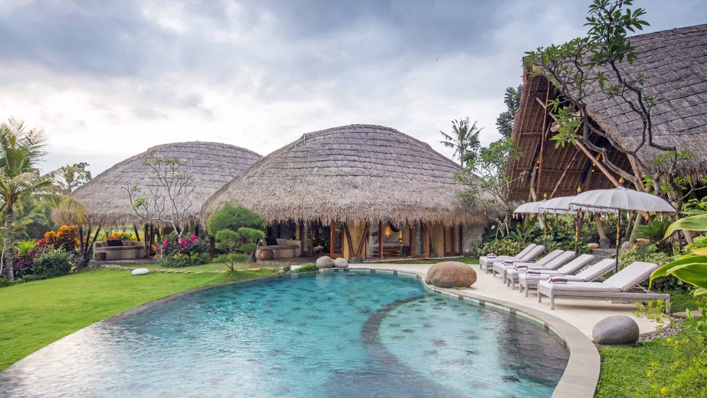 Luxurious Balinese-style thatched-roof villas with infinity pool, lounge chairs, and tropical gardens under a cloudy sky. - Bali Villas