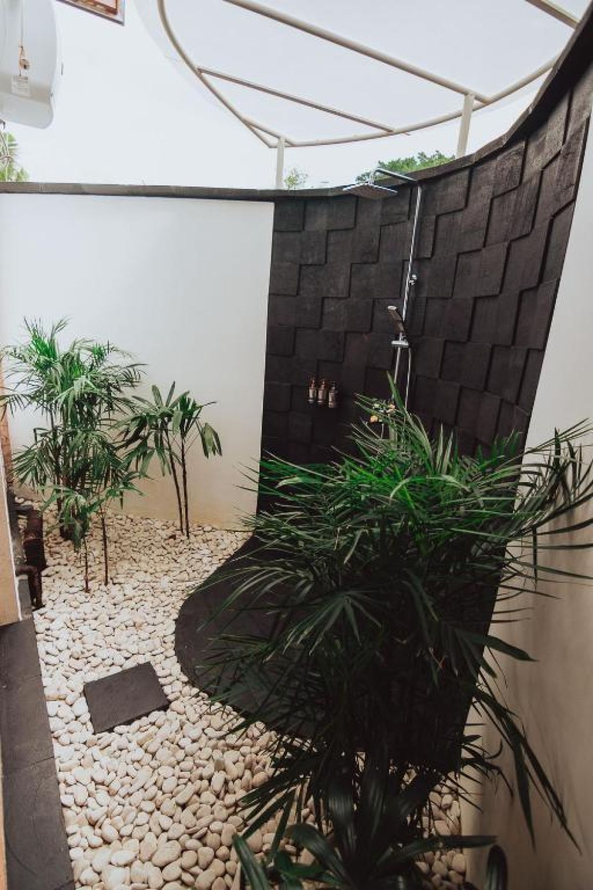 Modern outdoor shower with black pebble walls, tropical plants, hanging towels, and a sleek white sink. - Bali Villas