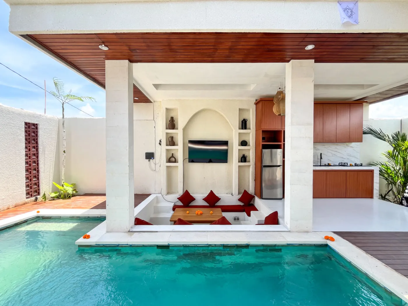 Luxurious Balinese villa outdoor pavilion with red cushioned seating, wooden kitchenette, and turquoise infinity pool. - Bali Villas