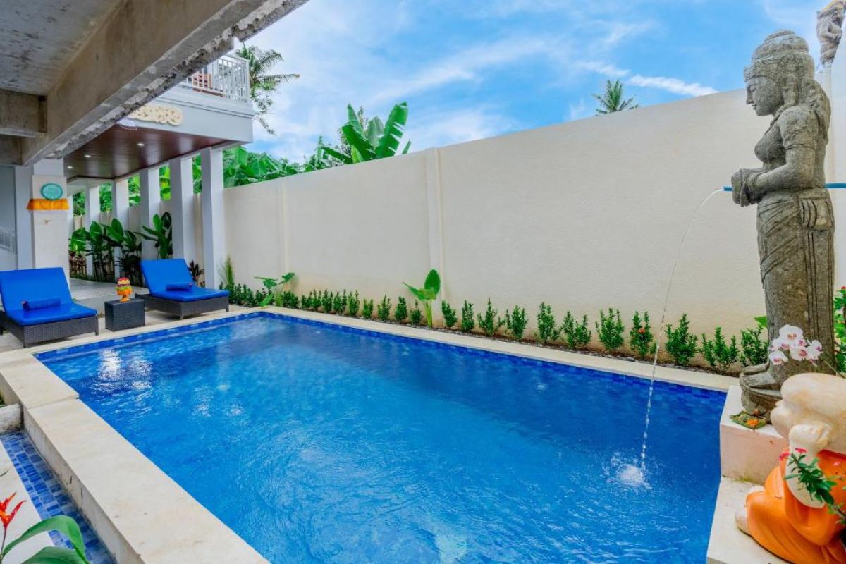 Serene tropical pool in a Balinese villa, featuring clear blue water, lounge chairs, lush plants, and stone statues of Hindu deities under a sunny sky. - Bali Villas