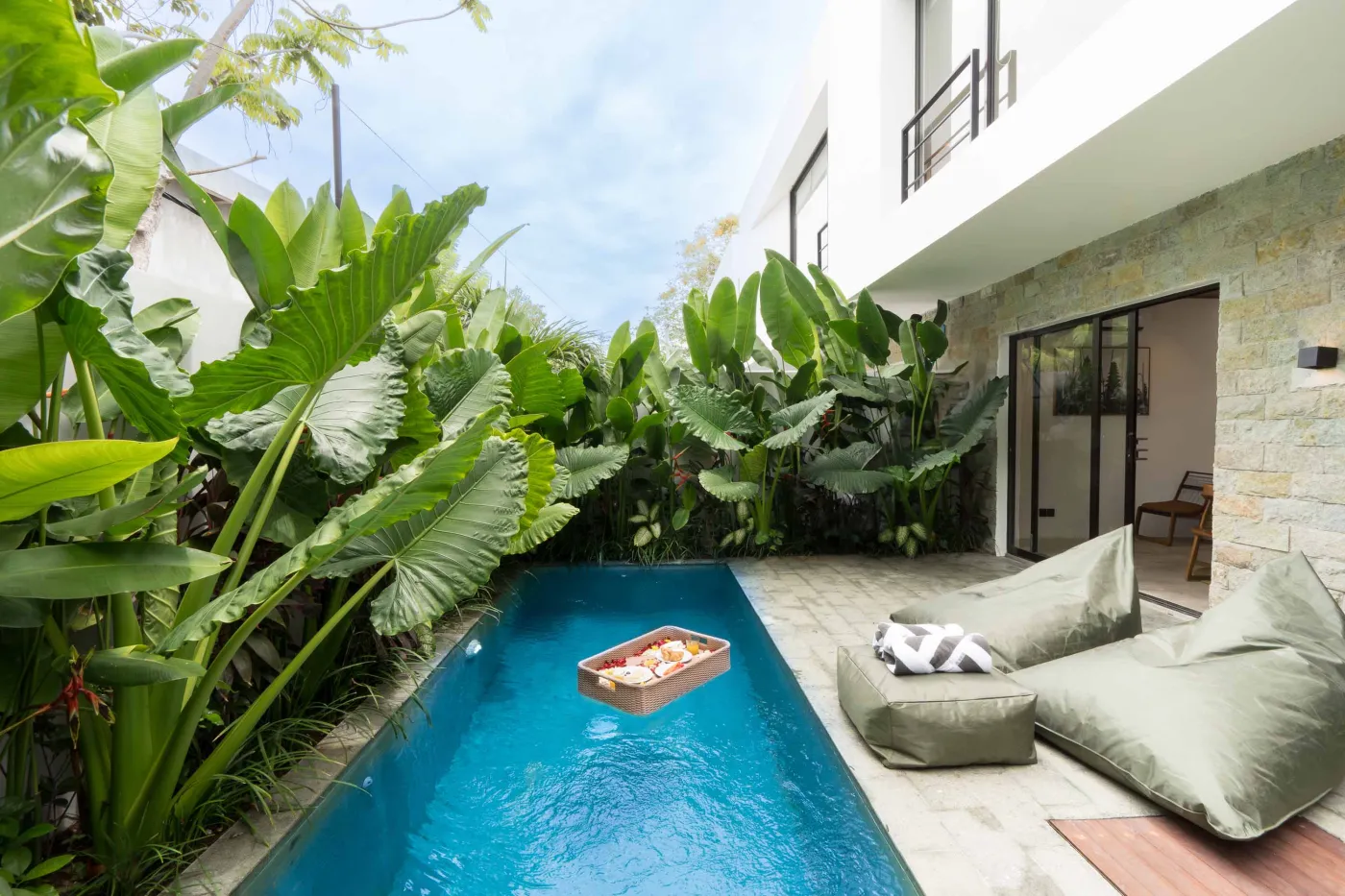 Serene tropical infinity pool in modern villa, surrounded by lush elephant ear plants, with floating fruit tray and cozy bean bags. - Bali Villas