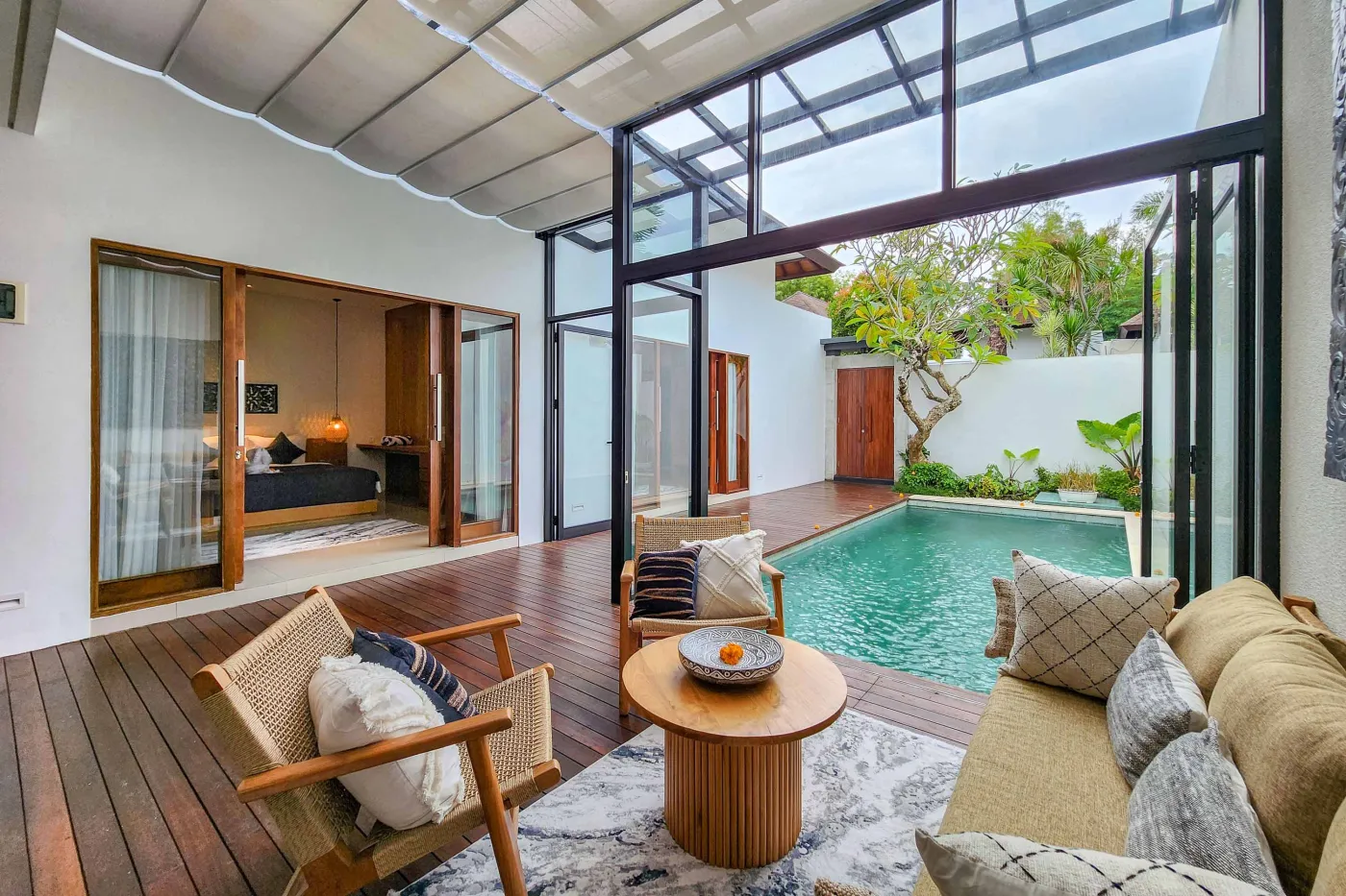 Luxurious tropical villa with private infinity pool, rattan lounge seating on wooden deck, and glass doors opening to a serene bedroom. - Bali Villas