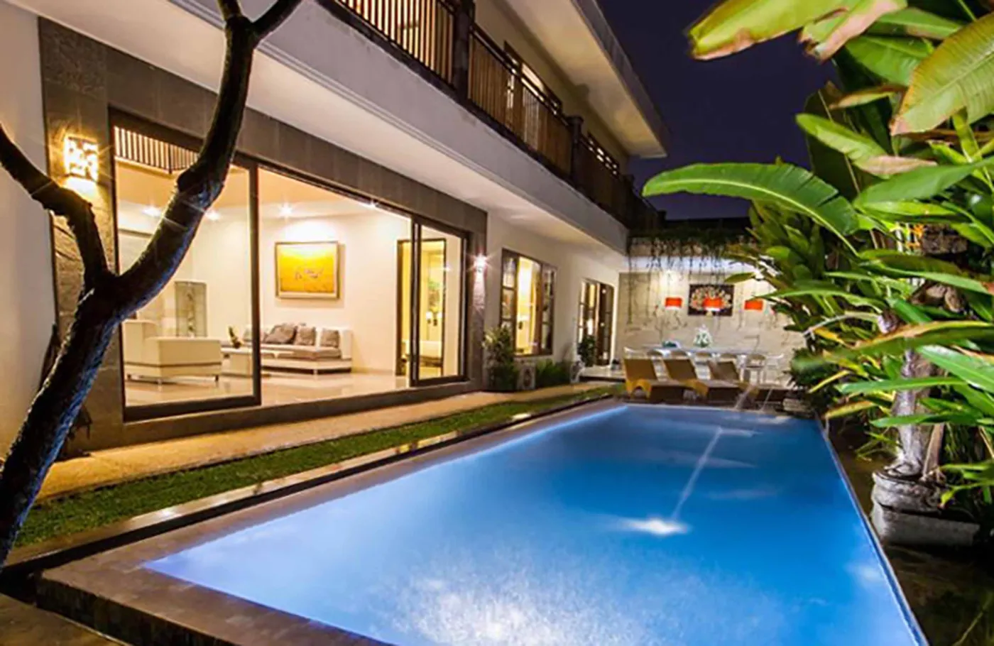 Luxury Bali villa at night with glowing blue infinity pool, modern glass-walled living area, and tropical plants. - Bali Villas
