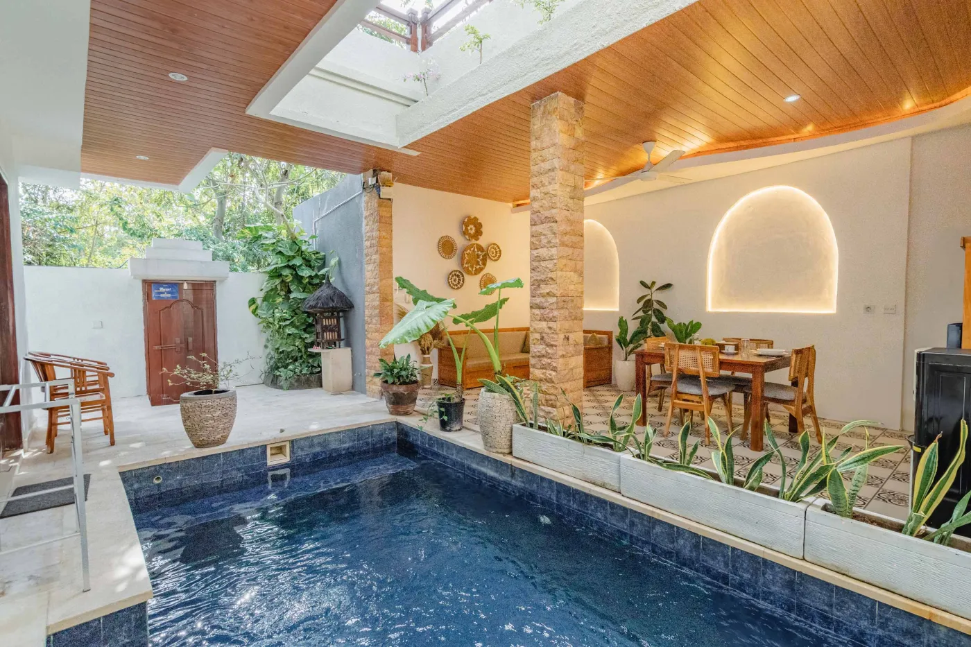 Serene tropical villa courtyard featuring a blue-tiled pool, lush plants, wooden dining area, and Balinese wall decor. - Bali Villas