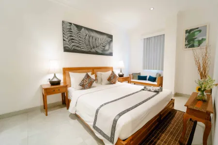 Cozy Balinese bedroom with wooden bed, white linens, fern wall art, and potted flowers.