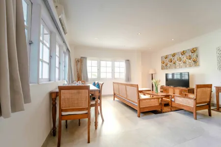 Spacious tropical villa interior with wooden dining table, rattan chairs, sofa, TV, and large open windows. - Bali Villas