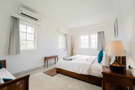 Spacious tropical bedroom with king bed, white linens, teak furniture, and large windows with sheer curtains.