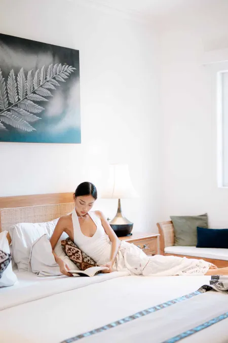 A young Asian woman lounging on a white bed in a serene bedroom, reading a book, with a large fern artwork on the wall.
