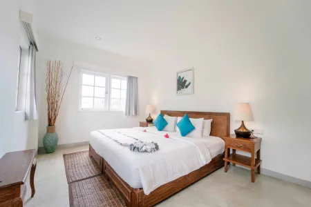 Bright tropical bedroom with wooden king bed, white linens, turquoise pillows, bedside lamps, and potted plant by large window. - Bali Villas