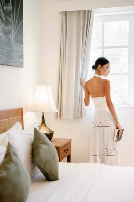 Elegant Asian woman in white embroidered maxi dress stands by window holding book in serene tropical bedroom with bed and lamp.