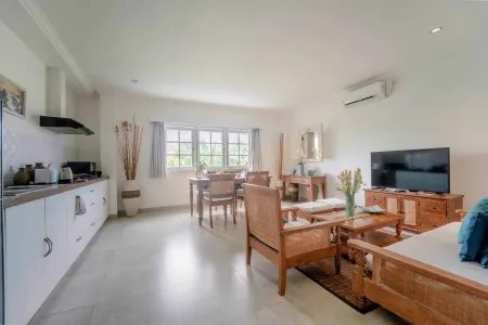 Spacious open-plan kitchen and living room in a tropical villa, with white cabinets, wooden dining table, teal sofa, large windows, and potted plants. - Bali Villas