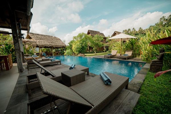 Serene Bali resort poolside with wicker lounge chairs, thatched pavilions, tropical gardens, and stone statues. - Bali Villas