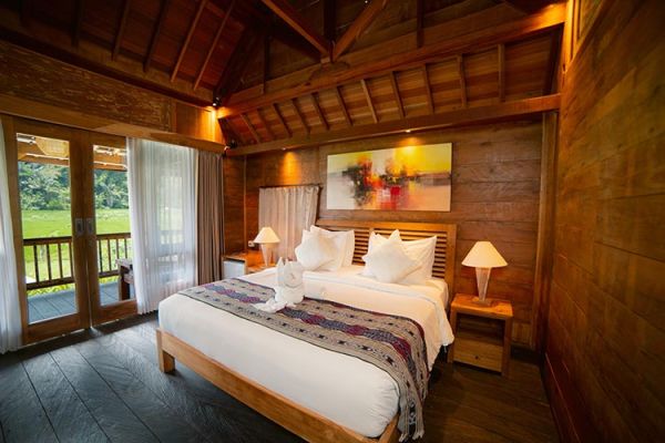 Luxurious tropical villa bedroom with wooden beams, king bed with white linens and colorful runner, bedside lamps, abstract wall art, and balcony doors opening to lush green views.