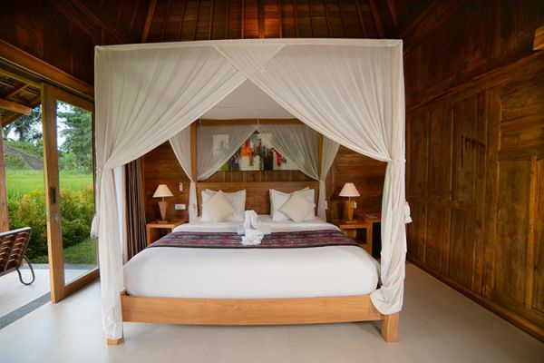 Elegant Balinese bedroom with wooden canopy bed, white mosquito net, and views of tropical greenery.