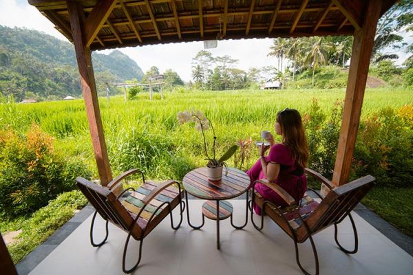 Woman in purple dress sips tea at wooden pavilion table with orchids, overlooking lush Bali rice terraces and mountains. - Bali Villas